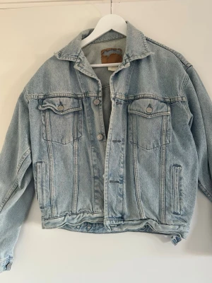  Ljusblå jeansjacka - Säljer en ljusblå jeansjacka med klassisk krage, två bröstfickor med lock och knappar samt två sidofickor. Jackan har en avslappnad passform och snygga sömdetaljer. 
