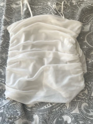 Vit mesh bandeau topp - Säljer en vit bandeau topp med tunna axelband och draperad mesh över hela toppen. Toppen är axelbandslös i stilen och har en tight passform som ger en trendig look. Perfekt att styla med jeans eller kjol för en cool outfit.
