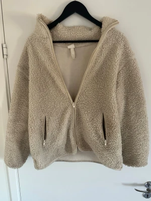 Beige teddyjacka med dragkedja - Mysig beige teddyjacka med fluffig struktur och två snedställda fickor med dragkedja framtill. Jackan har hög krage och hel dragkedja.