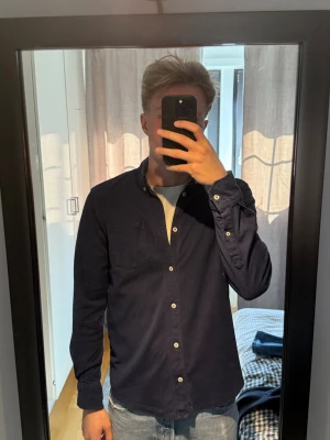 Mörkblå skjorta med knappar från Denham - Säljer en stilren mörkblå skjorta med vita knappar och klassisk krage. Skjortan har lång ärm och bröstficka, perfekt till jeans eller chinos. Materialet känns som bomull och passformen är normal. Skicket är bra och skjortan finns inte heller att köpa! 