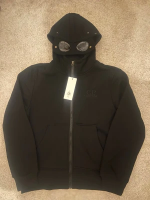 C.P. Company hoodie googles - Säljer en svart hoodie från C.P. Company med ikoniska inbyggda glasögon i huvan. Hoodien har dragkedja framtill, två stora fickor och diskret logga på bröstet. Tillverkad i mjukt material som känns skönt mot huden. Perfekt för dig som vill sticka ut med en unik detalj.
