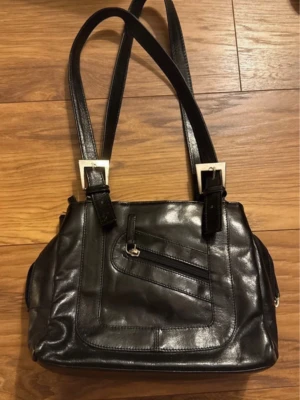 Svart axelremsväska i skinn - Snygg svart handväska i glansigt skinn med dubbla axelremmar och silverfärgade detaljer. Väskan har en framficka med dragkedja och ytterligare en ficka på baksidan. Perfekt storlek för det viktigaste och ger en stilren look. Många fickor, mycket rymlig! Tecken på användning! Hör av er vid frågor!