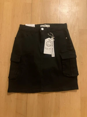 Svart cargo/jeanskjol stretch helt NY från Hello Miss - Snygg svart cargo/jeans kjol med stretch från Hello Miss med två stora sidofickor och bälteshällor i midjan. Kjolen har en rak passform och stängs med knapp och dragkedja framtill. Perfekt för en cool och avslappnad stil. Storlek 38/M perfekt till sommaren med en cool stretchig kjol