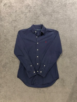 Marinblå skjorta från Ralph Lauren - Klassisk marinblå skjorta från Ralph Lauren med slim fit-passform. Skjortan har långa ärmar, vita knappar och den ikoniska röda logotypen broderad på bröstet. Tillverkad i mjuk bomull och perfekt för en stilren look.
