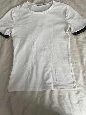 Vit t-shirt med svarta detaljer - Vit t-shirt med rund hals och korta ärmar. Ärmsluten har svarta glittriga detaljer som ger en snygg kontrast. Tillverkad i mjukt bomullsmaterial för skön känsla. Perfekt till jeans eller kjol för en enkel men trendig look.