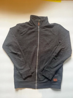 Mörkgrå ziptröja från Blend - Mörkgrå ziptröja från Blend med hög krage och hel dragkedja framtill. Tröjan har ribbade muddar vid ärmslut och nederkant samt en liten läderdetalj vid dragkedjan och på nederkanten. Tillverkad i mjukt material för en skön känsla.