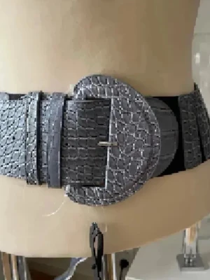 Brett grått bälte med strukturspänne - Snyggt brett bälte i grått med reptilmönstrad yta och stort, matchande spänne. Bältet har elastisk baksida för skön passform och ger en cool touch till din outfit. Perfekt för att markera midjan över klänning eller skjorta.Ny/aldrig använd 