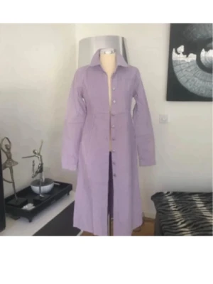 Ljuslila långklänning/kappa/kumono med knappar - Snygg ljuslila långklänning med krage och knappar hela vägen framtill. Klänningen har långa ärmar och markerad midja för en feminin siluett. Perfekt för dig som vill ha en stilren och trendig look. Manchester 