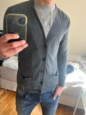 Mörkgrå tunn kofta med knappar - Snygg mörkgrå kofta med v-ringning och knäppning framtill. Modellen är tunn och har två fickor framtill samt ribbade muddar vid ärmslut och nederkant. Perfekt att slänga över en t-shirt för en clean look.