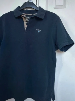 Svart pikétröja från Barbour - Klassisk svart pikétröja från Barbour med korta ärmar och krage. Tröjan har en liten logga broderad på bröstet och knappslå med diskret rutigt tyg på insidan. Perfekt till både jeans och shorts.