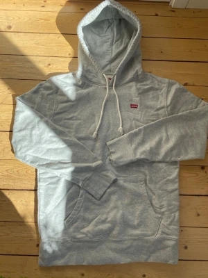 Grå hoodie från Levi's med huva - Snygg ljusgrå hoodie från Levi's med klassisk huva och vita dragsnören. Tröjan har en liten röd Levi's-logga på bröstet och en stor magficka framtill. Tillverkad i mjukt bomullsmaterial och har ribbade muddar vid ärmslut och nederkant.