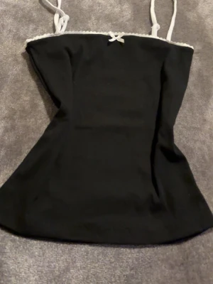 Svart linnetopp med vit spetskant - Snygg svart linnetopp med tunna axelband och vit spetskant upptill. Dekorerad med en liten rosett framtill för en söt touch. Perfekt att matcha med jeans eller kjol för en trendig look.