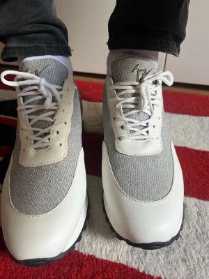 Gråvita sneakers från Giuseppe Zanotti modell Jimmy running  - Snygga sneakers i grått tyg och vitt läder från Giuseppe Zanotti. Skorna har vita snören, svart sula och en stilren siluett med låg profil. Perfekta för dig som gillar en clean och modern look med exklusiv känsla. Skorna är också helt nya. Pris från butik 7400kr mitt pris 5000kr