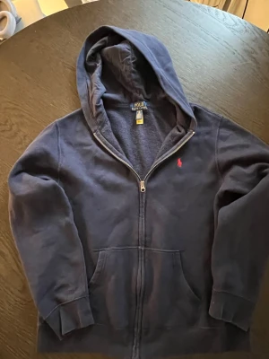 Mörkblå hoodie från Polo Ralph Lauren - Mörkblå hoodie från Polo Ralph Lauren med dragkedja framtill och klassisk röd logga broderad på bröstet. Hoodien har känguruficka och mjukt material, samt en stor huva. Perfekt för en avslappnad och stilren 