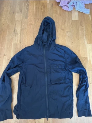 Svart vindjacka med huva - Snygg svart vindjacka med huva och dragkedja framtill. Jackan har en bröstficka med knappstängning och två sidofickor. Tillverkad i lätt och tunt material som passar perfekt för blåsiga dagar. Enkel och stilren design utan synligt märke.