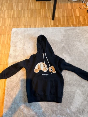 Palm Angels hoodie  - Säljer en svart hoodie från Palm Angels med en stor björn-illustration på bröstet och vit text under. Hoodien har huva med vita dragsnören och en klassisk magficka framtill. Perfekt för dig som gillar streetwear och vill sticka ut.