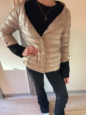 Herno jacka - Snygg beige dunjacka från Herno med tryckknappar framtill. Jackan har quiltad design, smal passform och diskreta fickor i sidorna. Modellen heter Elsa. Ärmarna är trekvarts längd och perfekt för nu till våren och sommaren och enkel att styla. IT 40 = EU 36/XS-S. Vid snabb affär är priset diskuterbart. Jackan är köpt i butik.
