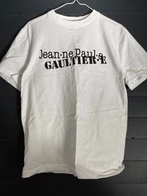 Vit Jean Paul Gaultier t-shirt - Snygg vit t-shirt merch med svart tryck framtill. Klassisk passform och rund halsringning. Perfekt för dig som gillar stilrena plagg med en twist. Tillverkad i mjuk bomull för skön känsla hela dagen.