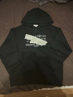 Maison Margiela svart hoodie - Svart hoodie från Maison Margiela med vit tryckt logga och siffror på bröstet. Klassisk passform med huva och långa ärmar. Tillverkad i mjukt bomullsmaterial, perfekt för lager-på-lager under kyliga dagar.