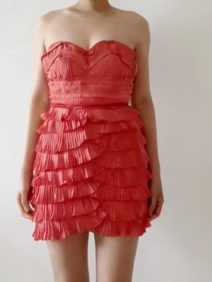 Axelbandslös kort volangklänning - Säljer en axelbandslös kort klänning i korallrosa med flera lager volanger och plisserade detaljer. Klänningen har en hjärtformad överdel och är perfekt för dig som gillar att sticka ut med färg och struktur. Materialet känns lätt och luftigt.