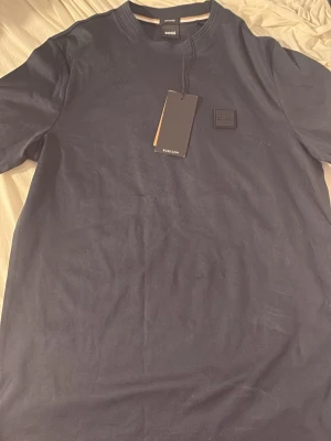 Mörkblå t-shirt från BOSS - Snygg mörkblå t-shirt från BOSS med rund halsringning och diskret logopatch på bröstet. Tillverkad i merceriserad bomull för extra mjuk känsla och stilren look. Perfekt för dig som gillar minimalistisk design och premiumkänsla.