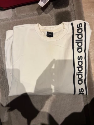 Vit sweatshirt från Adidas med logga - Säljer en vit sweatshirt från Adidas med svart logga och text längs ena sidan framtill. Tröjan har rund halsringning och är tillverkad i mjukt bomullsmaterial. Perfekt för en sportig och avslappnad stil.