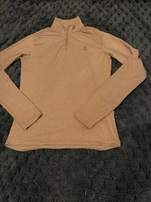 Beige sporttröja med halvzip - Sportig beige långärmad tröja med halvzip och hög krage. Tillverkad i ett stretchigt funktionsmaterial som andas, perfekt för träning eller utomhusaktiviteter. Diskret logga på bröstet och stilren design.