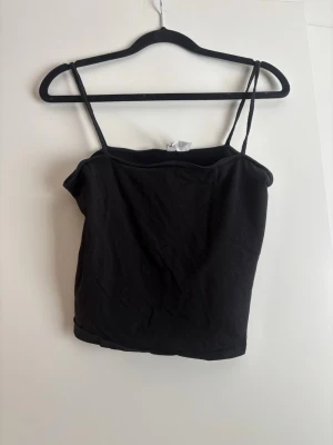 Svart linnetopp med smala axelband - Enkel och stilren svart linnetopp med smala axelband. Perfekt att matcha med jeans eller kjol för en clean look. Mjuk och bekväm, lätt att bära till olika outfits. Den är cropped i passformen