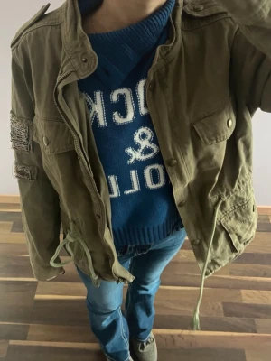 Olivgrön utilityjacka med detaljer - Snygg olivgrön utilityjacka med dragkedja och knappar framtill. Jackan har flera fickor, justerbar midja med snörning och dekorativ patch på ena ärmen. Tillverkad i ett slitstarkt bomullstyg som ger en avslappnad streetkänsla. nypris 999 kr. har en liten fläck på insidan (se bild) 