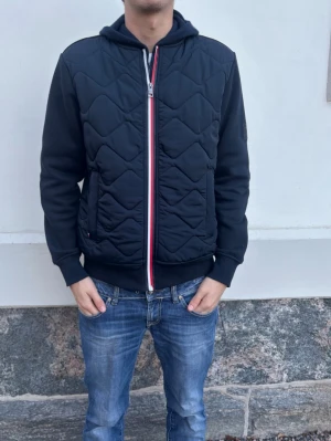 Tommy Hilfiger cardigan  - Svart quiltad bomberjacka från Tommy Hilfiger med huva och dragkedja framtill. Jackan har ribbade muddar, två fickor och en rödvit detalj längs dragkedjan. Liten logga på ärmen och sportig look.