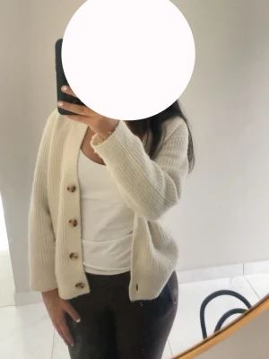 Crèmevit ribbstickad kofta med knappar - Mysig crémevit ribbstickad kofta med träknappar framtill. Koftan har en avslappnad passform, långa ärmar och är gjord i ett mjukt material som passar perfekt till lager på lager. Enkel och stilren design som funkar till det mesta. Använd 3 gånger.