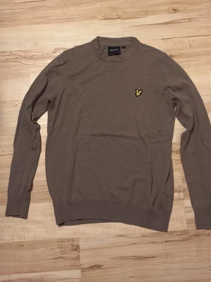 Beige stickad tröja Lyle & Scott - Snygg beige stickad tröja från Lyle & Scott med rund halsringning och den klassiska gula loggan broderad på bröstet. Tröjan har långa ärmar och ribbade muddar vid ärmslut och nederkant. Perfekt för lager på lager och stilrena outfits. Pris kan diskuteras