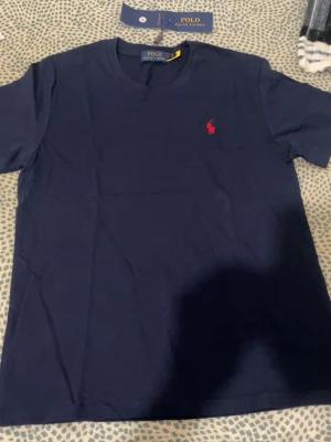 Marinblå t-shirt från Polo Ralph Lauren - Klassisk marinblå t-shirt från Polo Ralph Lauren med röd broderad logga på bröstet. T-shirten har rund halsringning och korta ärmar. Tillverkad i mjuk bomull för en skön känsla och enkel stil.