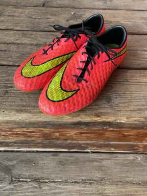 Nike hypervenom 1 elite fotbollsskor  - Säljer ett par röda Nike fotbollsskor med stora neongula och svarta swoosh-loggor. Skorna har snörning, låg profil och dobbar för spel på gräs. Ovandelen är i syntetmaterial med textur för bättre bollkontroll. Perfekta för dig som vill sticka ut på planen.