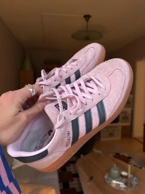 Adidas Handball Spezial rosa blå sneakers - Säljer ett par Adidas Handball Spezial sneakers i ljusrosa mocka med mörkblå ränder och gummisula. Klassisk och cool retro streetstyle med låg profil och snygga detaljer. Perfekta för dig som vill sticka ut med färg och ikonisk design. Använt ett par gånger bara för att testa men de är tyvärr inte min storlek!! (därav hälsulan som kan tas bort)