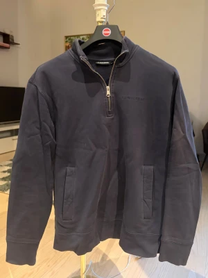 Mörkblå half-zip tröja J.Lindeberg - Mörkblå half zip från J.Lindeberg med en diskret broderad logga. Lite tjockare material så väldigt bekväm att bära. Gör sig snygg med både T-shirt och skjorta under. Tillverkad i mjukt bomullsmaterial för en avslappnad look. Passar S/M.