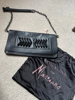Svart kuvertväska Elin Kling for Marciano - Stilren svart kuvertväska i läder med dekorativa detaljer framtill och en cool kedjerem. Väskan har ett unikt mönster på framsidan och kommer med en matchande dustbag. Perfekt accessoar för att lyfta din outfit.