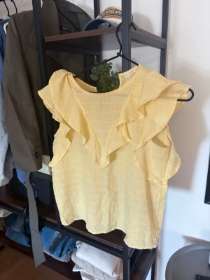 Gul ärmlös blus med volang - Söt gul blus med volangdetaljer runt halsringningen och axlarna. Blusen är ärmlös och har ett lätt rutmönster i tyget. Perfekt för en somrig och fräsch look.