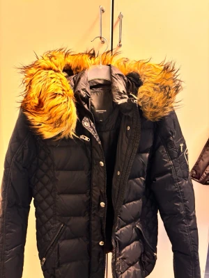 Svart rockandblue jacka med fuskpäls - Snygg svart äkta rockandblue med stor huva och fluffig fuskpäls i gult och brunt. Jackan har quiltade detaljer, dragkedjor på ärmarna och flera fickor framtill. Perfekt för kalla vinterdagar och ger en cool streetstil.