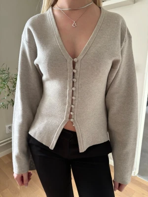 Beige cardigan med v-ringning - Trendig beige cardigan med djup v-ringning och dekorativa knappar framtill. Långärmad modell i mjukt stickat material som ger en clean och stilren look. Perfekt att matcha med både jeans och kjol för en avslappnad vibe.