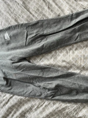 Gråa mjukisbyxor från Nike - Säljer ett par klassiska gråa mjukisbyxor från Nike med vit logga på låret. Byxorna har resår i midjan och vid bensluten för en skön passform. Tillverkade i mjukt bomullsmaterial som är perfekt för chill dagar eller träning.