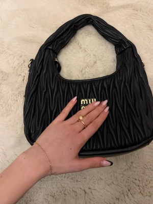 Svart handväska i läder - Säljer en svart handväska i quiltat läder med guldfärgad logga framtill. Väskan har en mjuk, rundad form och kort axelrem. Perfekt för dig som gillar lyxiga detaljer och trendig design.