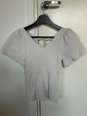 Ljusgrå ribbad topp med puffärm - Trendig ljusgrå topp med ribbad struktur och puffiga korta ärmar. Djup v-ringning både fram och bak samt en söt knytning i ryggen. Tillverkad i ett stretchigt, mjukt material som sitter snyggt på kroppen. Perfekt för dig som gillar en feminin och modern stil.