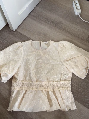 Krämvit blus med puffärm från Dobber - Supersöt krämvit blus från Dobber med puffiga korta ärmar och broderat blommönster. Blusen har rund halsringning, smockad midja och volang nedtill. Materialet är glansigt och ger en lyxig känsla. Perfekt för dig som gillar romantisk och trendig stil.