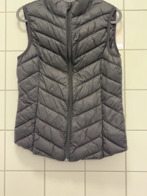 Mörkgrå dunväst med dragkedja - Snygg mörkgrå dunväst med hög krage och dragkedja framtill. Västen har ett quiltat mönster i diagonala ränder och är ärmlös. Perfekt att slänga över en hoodie eller tröja för extra värme. Materialet är lätt och smidigt, vilket gör den enkel att bära.