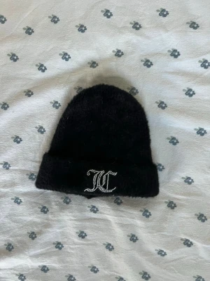 Svart fluffig mössa från Juicy Couture - Säljer en svart, fluffig mössa från juicy couture med uppvikt kant och glittrande JC-logga framtill i strass. Mössan har en enkel och stilren design som passar perfekt till kalla dagar. Materialet ser mjukt och mysigt ut. Aldrig använt. 