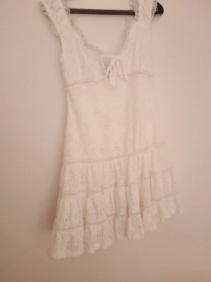 Vit spetsklänning med volang - Supersöt vit kortklänning i bomull med spetsdetaljer och volangkant. Klänningen har knyt framtill och breda axelband. Perfekt för sommarens alla tillfällen och ger en romantisk vibe.