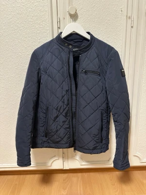 Mörkblå quiltad jacka från Replay - Snygg mörkblå quiltad jacka från Replay med dragkedja och ståkrage. Jackan har ett diskret rutmönster, tre fickor med dragkedja och justerbara detaljer vid midjan. Perfekt för dig som vill ha en stilren och modern look.