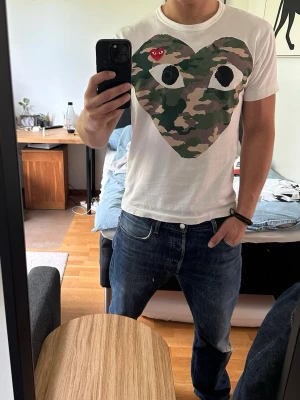 Vit Comme des Garçons Play t-shirt - Säljer en vit t-shirt från Comme des Garçons Play med stort hjärta i camouflage på bröstet och det klassiska röda hjärtat med ögon. T-shirten har rund hals och korta ärmar, perfekt för en clean och trendig look.