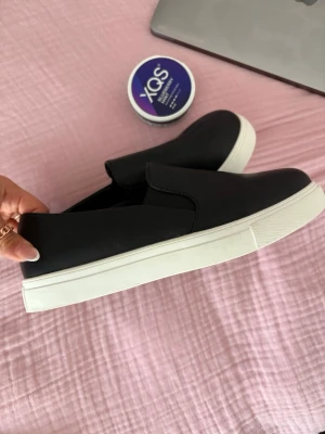 Svarta slip-on sneakers - Stilrena svarta slip-on sneakers med vit sula. Skorna har en minimalistisk design utan snörning och är tillverkade i ett slätt material som ger en clean look. Perfekta för dig som gillar enkel och modern stil.